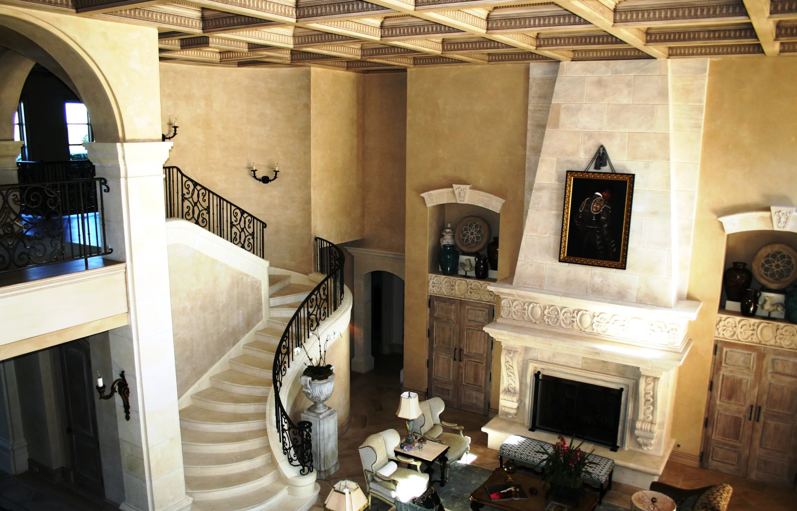 Luxury interior featuring a curved stone staircase, decorative wrought iron railings, coffered ceiling, ornate fireplace, and architectural millwork.